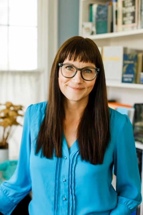 Author photo of Maggie Smith in a blue shirt and glasses and standing in front of a bookcase.