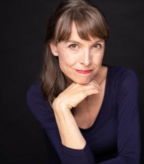 A head-and-shoulders portrait of author Nita Prose. She has light-colored hair with bangs, styled in a slightly off-center part, and is wearing a dark blue long-sleeved top. Her chin rests on her hand, and she is looking directly at the camera with a slight smile. The background is a solid black color, creating a high-contrast image that focuses attention on her face.