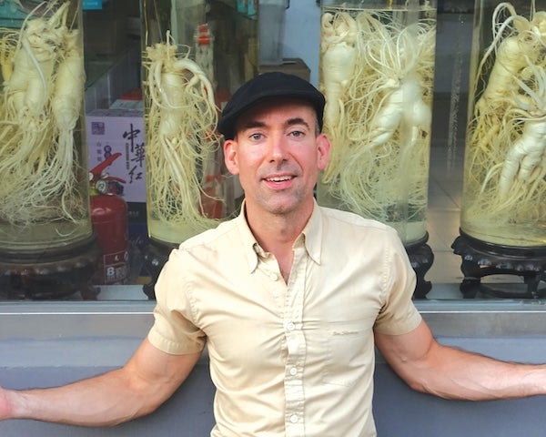 Author photo of Craig Thompson. Craig is outside in front of glass cases of ginseng, wearing a short-sleeved button-up shirt and a pageboy hat.