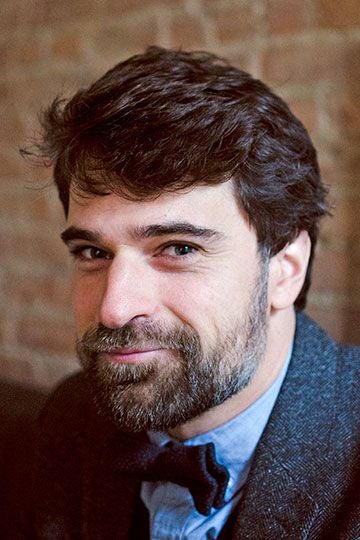 Author photo of Dane Huckelbridge, who is in front of a brick wall which is out of focus, wearing a suit shirt and blazer and bowtie.