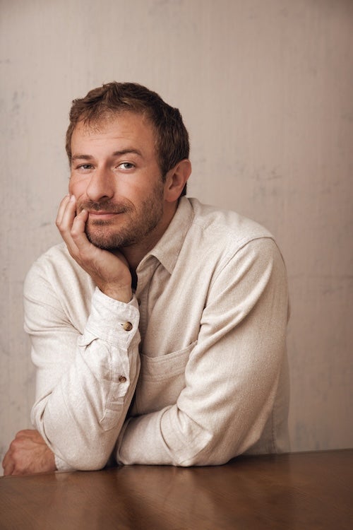 Author photo for Gabe Henry. Gave is leaning against a wooden table with his right hand cupped under his chin, looking directly at the camera, and in front of a cream background.