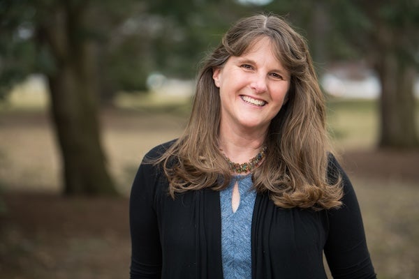 Author photo of Kate Messner. Kate stands outside in front of a field and trees, which are blurry in the background. She's wearing a black cardigan over a blue shirt, and she is smiling at the camera.