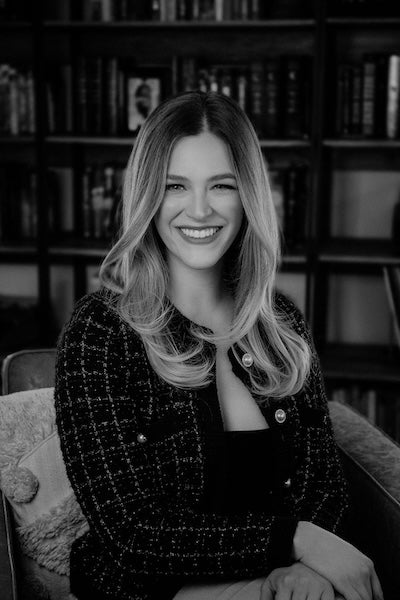 Author photo for Kelsey Impicciche. Kelsey sits in a chair and in front of a bookshelf, and the image is in black and white.