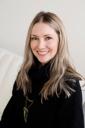 A head-and-shoulders portrait of author Lauren J. A. Bear. She has long blonde hair and is wearing a black turtleneck sweater and a long necklace with a green pendant. She is smiling and looking directly at the camera. The background is a plain, light-colored wall.