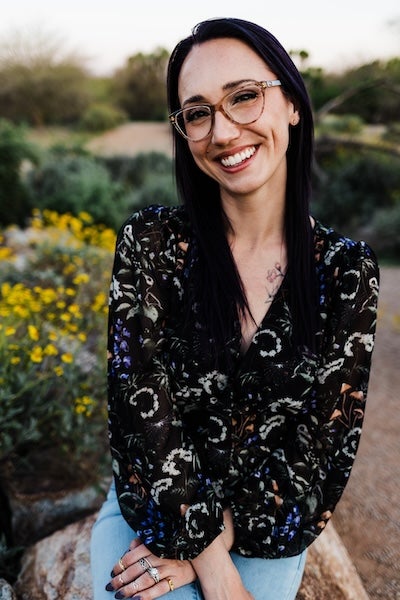 Author photo for Maxym M. Martineau outside standing in front of wildflowers, wearing glasses and smiling with her teeth, looking at the camera.