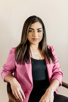 A portrait of author Neely Tubati Alexander. She has long, dark hair with lighter highlights and is wearing a bright pink blazer over a black top. She is seated, looking directly at the camera with a neutral expression. She is wearing a delicate necklace and rings on her fingers. The background is a plain, light beige wall. The lighting is soft and even.