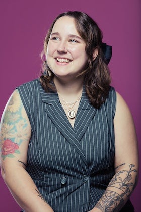 A portrait of Kimm Topping. Kimm has shoulder-length wavy brown hair with a dark blue bow tied in it. They are smiling and looking to the right. Kimm is wearing a grey striped vest over a lighter grey shirt and a necklace with a round pendant. Their arms are visible, revealing tattoos on both forearms. The background is a solid bright pink.