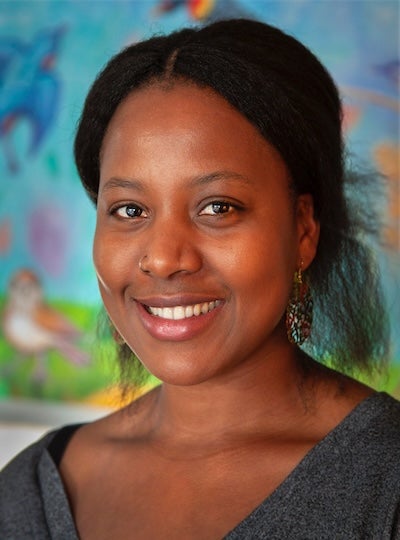 Author photo of Iryn Tushabe. Iryn stands in front of a painted mural, which is blurry, and smiles at the camera.