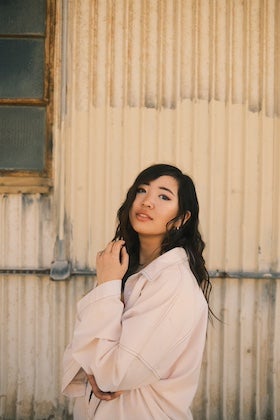 A portrait of author Jemimah Wei. She has long, wavy dark hair and is wearing a light pink, collared shirt. She is standing indoors, looking slightly upwards and to her right, and her right hand is gently touching her face. The background is a textured, light-colored wall with a window visible on the left side.