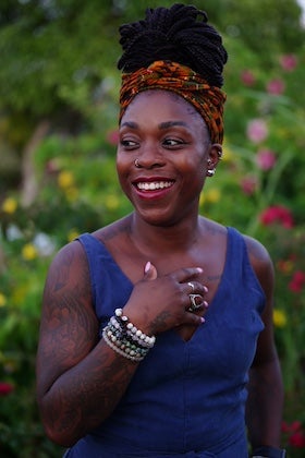 A portrait of author Tamla T. Young from the chest up. She appears to be a Black woman with dark, styled hair wrapped in a vibrant orange and brown patterned headwrap. She is wearing a sleeveless blue top and several beaded bracelets on her left wrist. Her left hand is gently placed near her chest. She has a nose ring and earrings, and she is smiling broadly, looking off to the side. The background is a soft blur of green and colorful flowers, suggesting an outdoor setting.