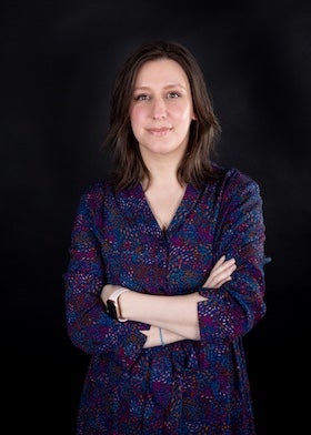 A portrait of YA author Sarah Daniels. She has shoulder-length brown hair and is wearing a dark blue dress with a colorful pattern. She is standing with her arms crossed and looking directly at the camera with a slight smile. She is wearing a watch on her left wrist and a thin bracelet on her right wrist. The background is a solid black color.