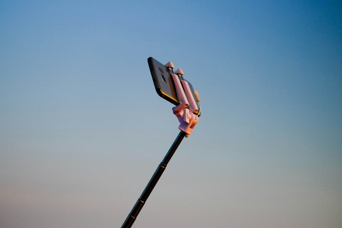 A cell phone, likely a smartphone, is mounted on a selfie stick, extending upwards against a clear, gradient blue sky. The sky is lighter blue towards the bottom, transitioning to a darker, deeper blue at the top. The phone is held in a light pink or purple holder, and the stick itself is dark. The sun appears to be hitting the phone and holder from the side, creating a slight glint and highlighting their form against the smooth background of the sky.