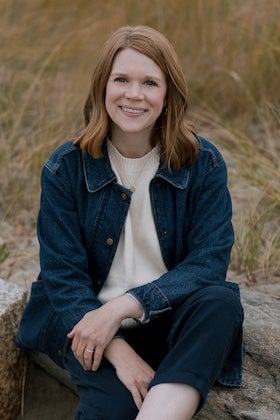 A headshot of Samantha Browning Shea, a young woman with reddish-brown, wavy hair, smiling gently at the viewer. She is seated outdoors, wearing a cream-colored sweater layered under a dark blue denim jacket. In the background, out of focus, are dry, golden grasses and what appear to be rocks or sand, suggesting a beach or dunes environment.