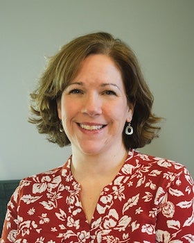 A headshot of Rhonda DeChambeau, a woman with short, wavy brown hair, smiling warmly. She is wearing a rust-red colored blouse with a white floral or leaf pattern and a v-neckline. She is positioned slightly to the right, looking directly at the viewer, against a plain, light grey background.