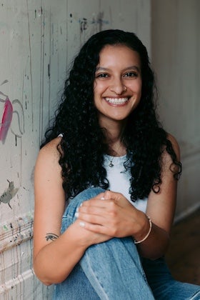 A headshot of Deena ElGenaidi, a young woman with long, dark, curly hair and warm brown skin, smiling broadly at the viewer. She has a nose ring and is wearing a simple white sleeveless top. She is seated, with her knees drawn up, holding her hands clasped. The background is a textured light wall with some faint marks and what appears to be abstract paint streaks.