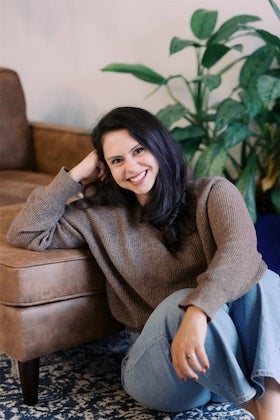 A headshot of Hannah Rosenberg, a woman with dark hair and a warm smile, seated on the floor. She is leaning against a brown leather couch or armchair, with her arm resting on it, and looking towards the viewer. She is wearing a cozy, brown knitted sweater and light-wash blue jeans. In the background, a large green plant is visible.
