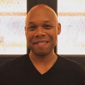 A headshot of Tre Johnson, a Black man with a shaved head, smiling warmly and looking directly at the viewer. He has a nose ring on his left nostril. He is wearing a black v-neck shirt. The background is blurred, showing what appears to be horizontal blinds or a window with horizontal panes.