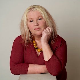 A headshot of Hazell Ward, a woman with long, light blonde hair with some pinkish highlights, looking directly at the viewer with a thoughtful expression. She has her right hand resting under her chin. She is wearing a deep red, v-neck top and a chunky, amber-colored beaded necklace. The background is a plain, light grey.