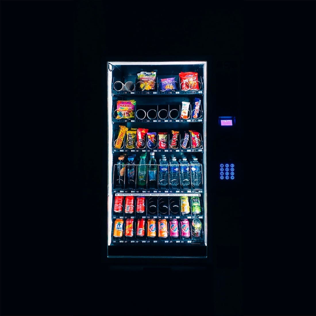 A vending machine lit up to display sodas, waters, and various candy bars. Everything around the machine is totally black, giving it a mystical glow.