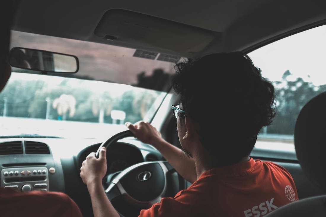 Taken from the backseat in a car, it features a curly-haired masculine person behind the wheel of the car.